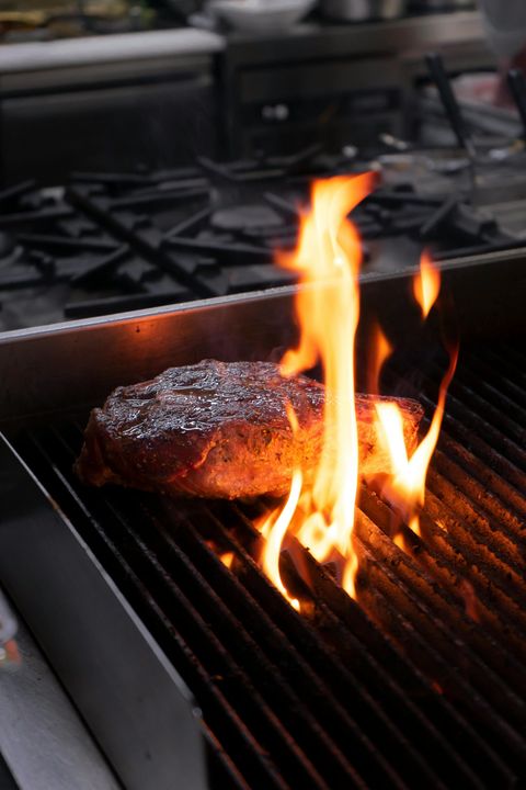 A raw steak sits on a hot grill grate with bright orange flames rising up around the meat.