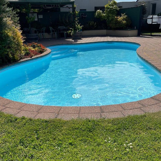 A kidney-shaped swimming pool with bright blue water, surrounded by paved stone decking and a lush green lawn.