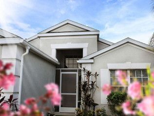 Beige house with white trim and a screen door, pink flowers in the foreground against a bright blue sky.