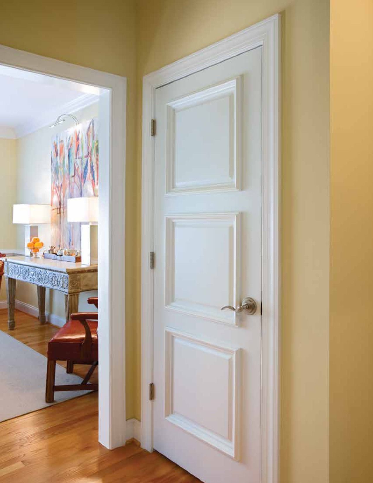White door with three recessed panels in a yellow-walled hallway, leading to a room with artwork and furniture.