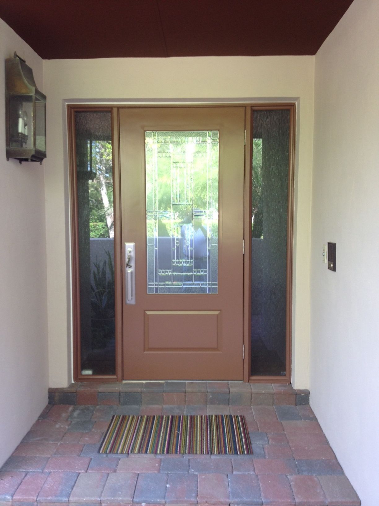 Tan front door with sidelights, doormat, and brick walkway.