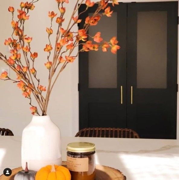 White vase with orange flowers on table with pumpkins and black doors.