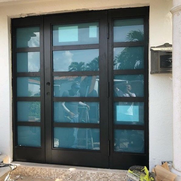 Black framed front door with glass panels, reflections of people, building exterior.