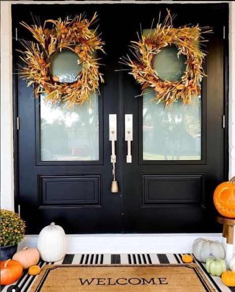 Black double doors with wheat wreaths, pumpkins, and a welcome mat, creating a fall-themed entryway.