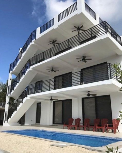 White multi-story building with black railings, balconies, and a pool. Red chairs in front.