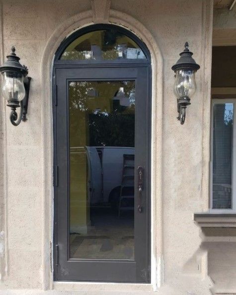 Dark gray front door with arched window, surrounded by beige stucco and two wall sconces.