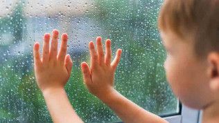 Child with hands pressed against a window covered in raindrops, looking outside.