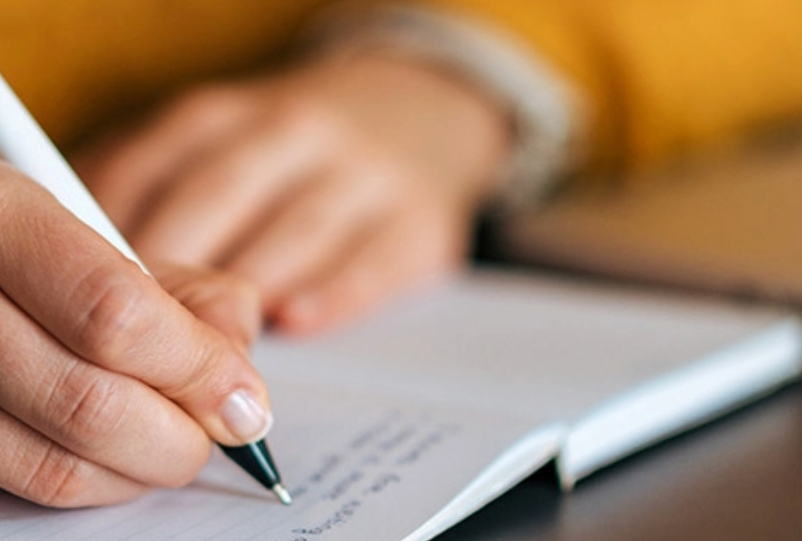 Eine Person schreibt mit einem weißen Stift in ein Notizbuch. Nahaufnahme. Im Hintergrund ein gelber Pullover.