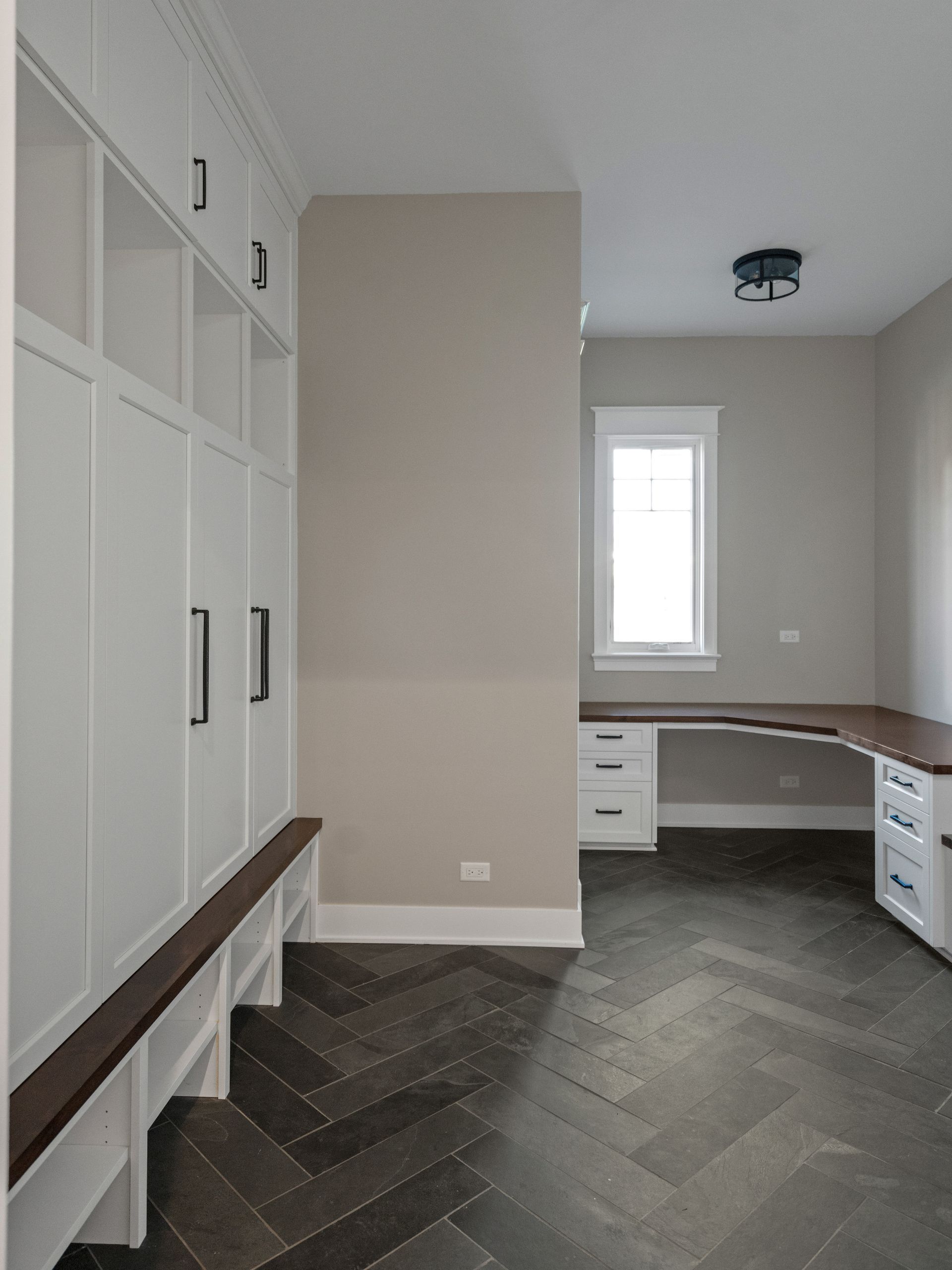 A room with a herringbone floor , white cabinets , a desk and a window.