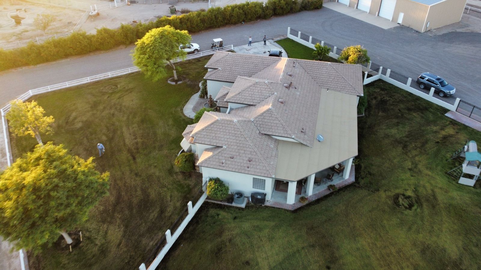 Aerial view of a beige house with a brown roof on a grassy lot, surrounded by a white fence.
