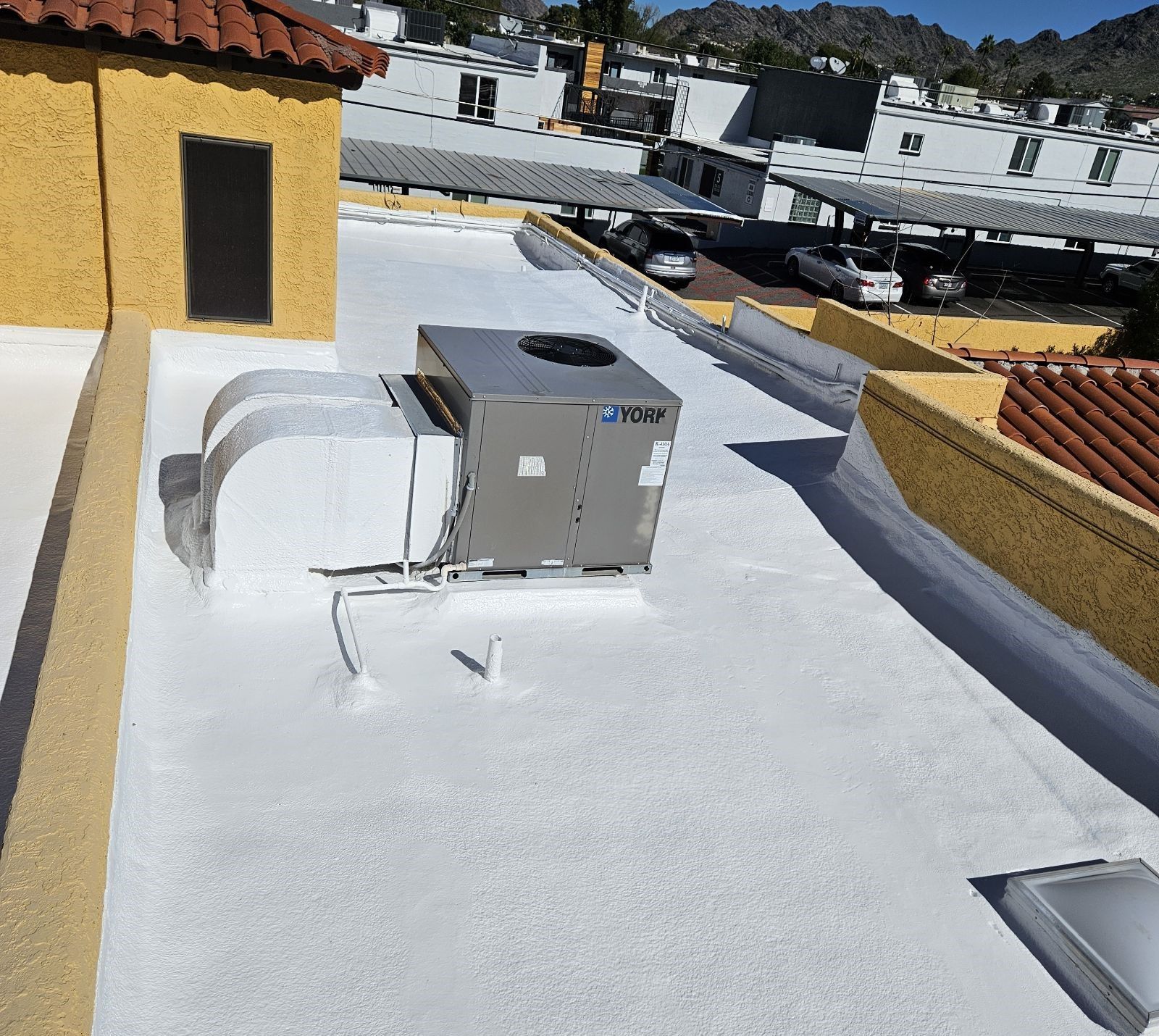 White rooftop with HVAC unit, and ventilation ducts. Sunny day with other buildings in the distance.