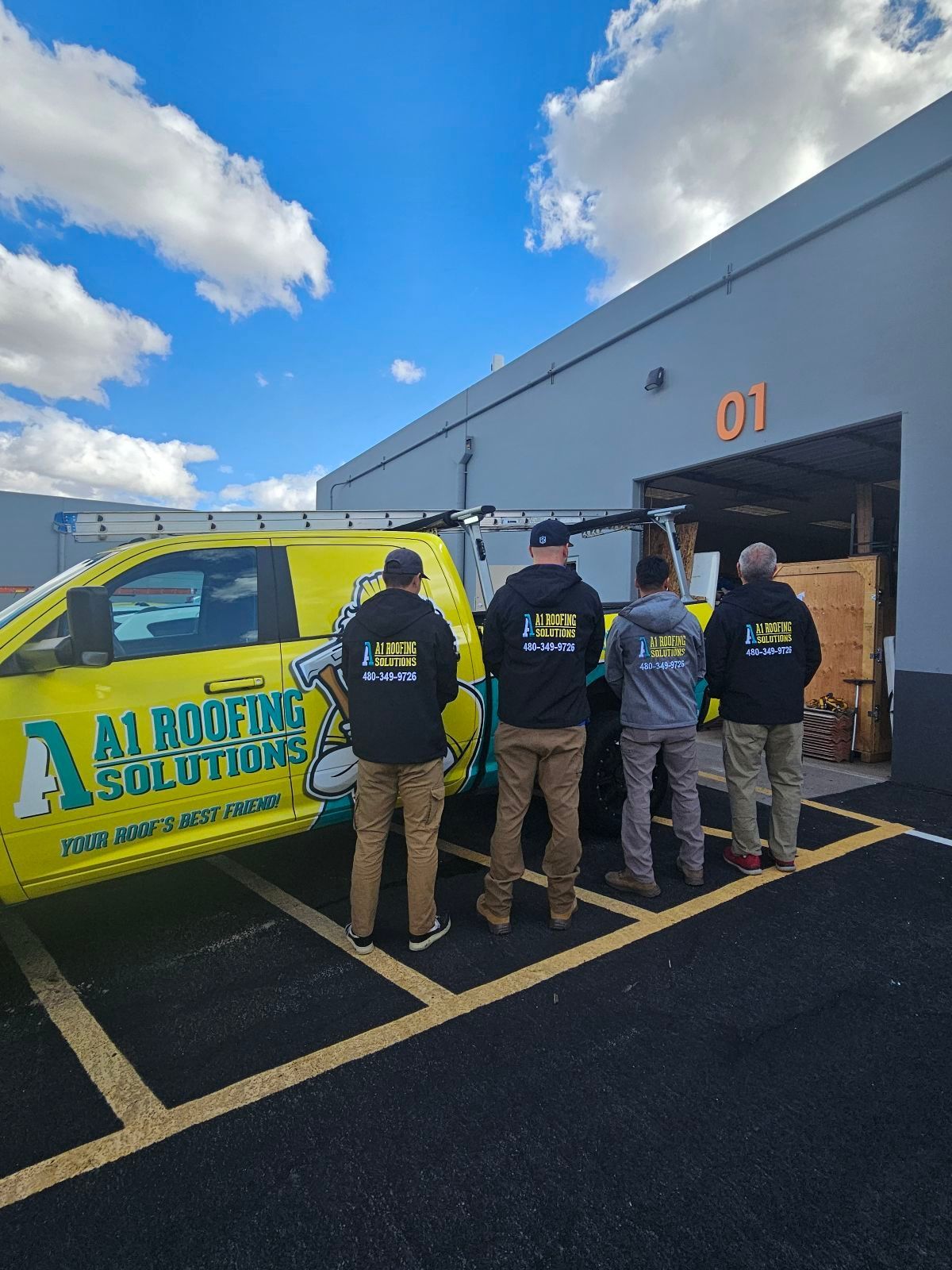 Four people in workwear standing in front of a yellow roofing truck and a building with 