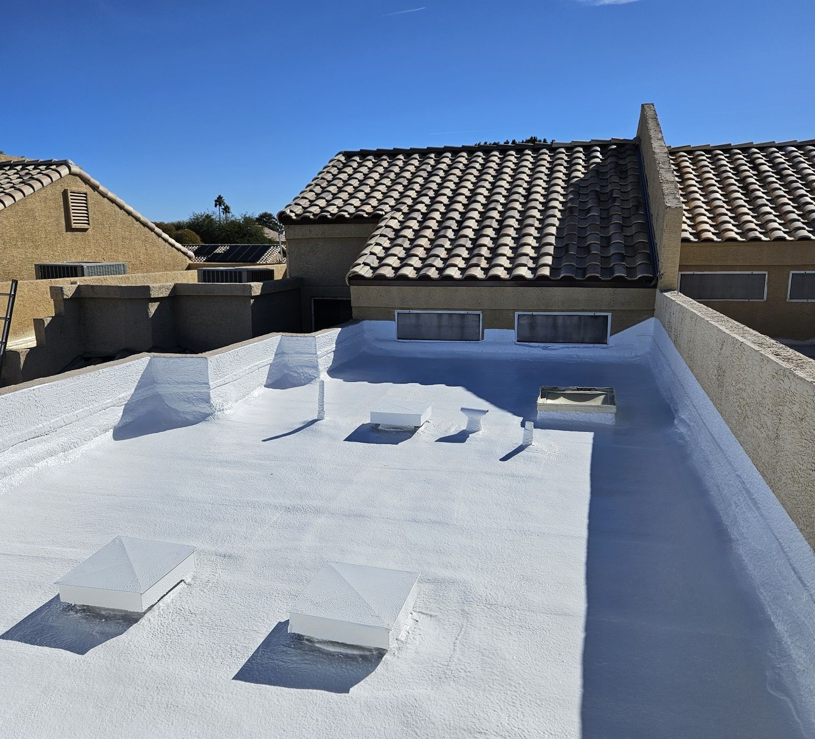 White roof coating on a flat roof with air vents, adjacent to buildings with tile roofs and a clear blue sky.