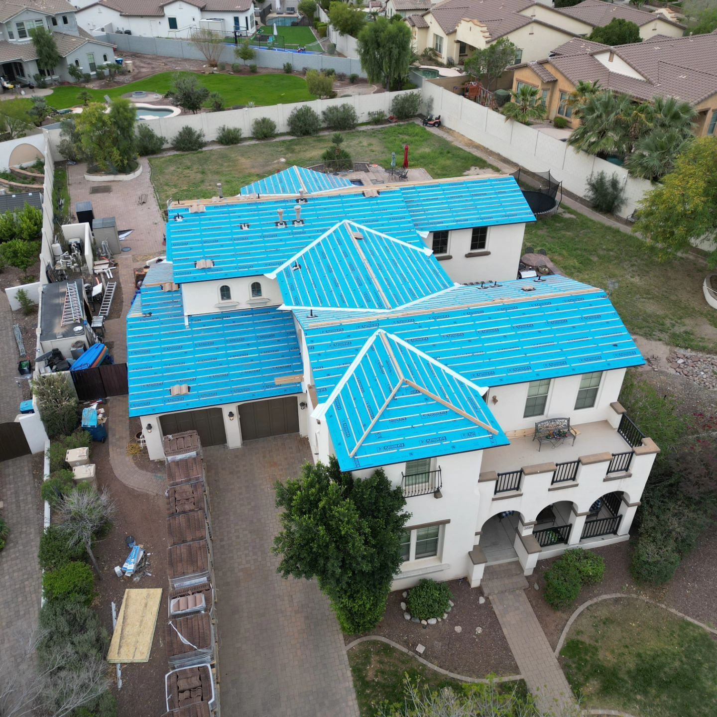 An aerial view of a two-story house with blue tiled roof and white stucco exterior in a neighborhood.