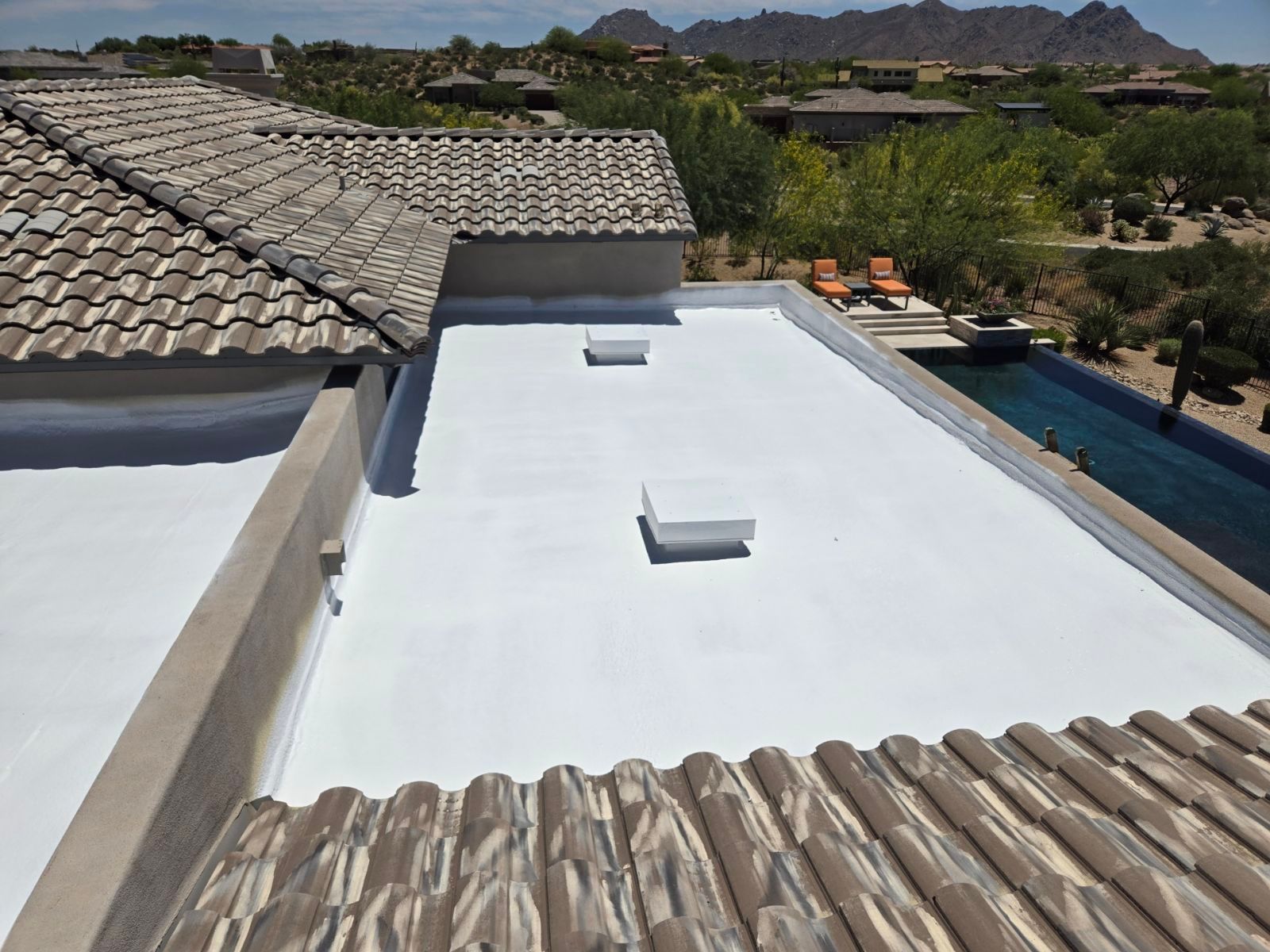 Flat white roof with skylights, surrounded by a tile roof, near a swimming pool and mountains.