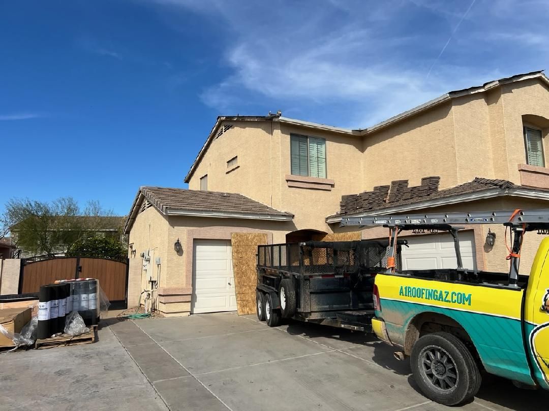 A tan stucco house with a truck and trailer loaded with roofing materials under a blue sky.