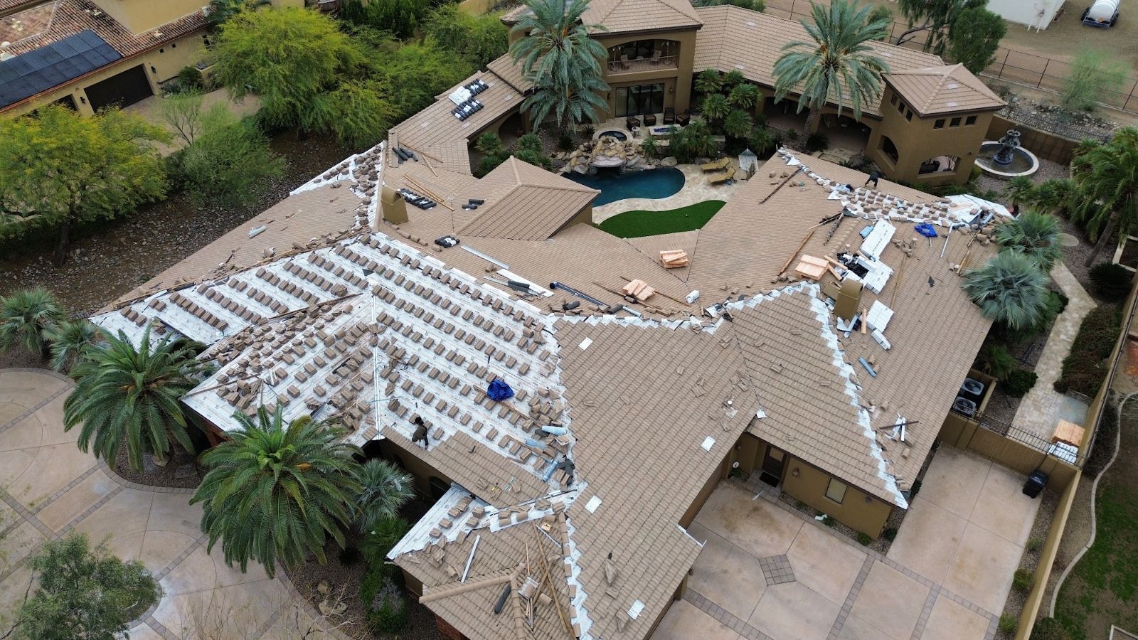 Aerial view of a home with roof tiles being replaced; pool, palm trees, and work supplies are visible.