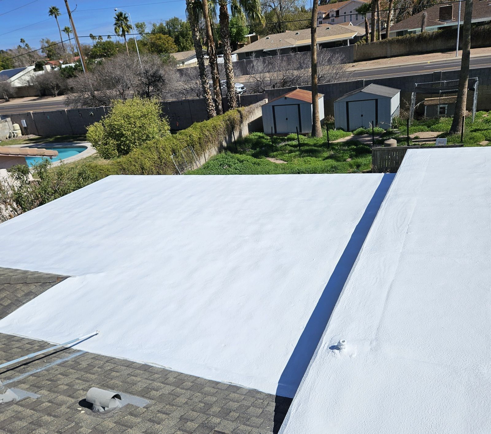 Flat roof partially covered in white coating; view overlooking yard with pool and sheds.