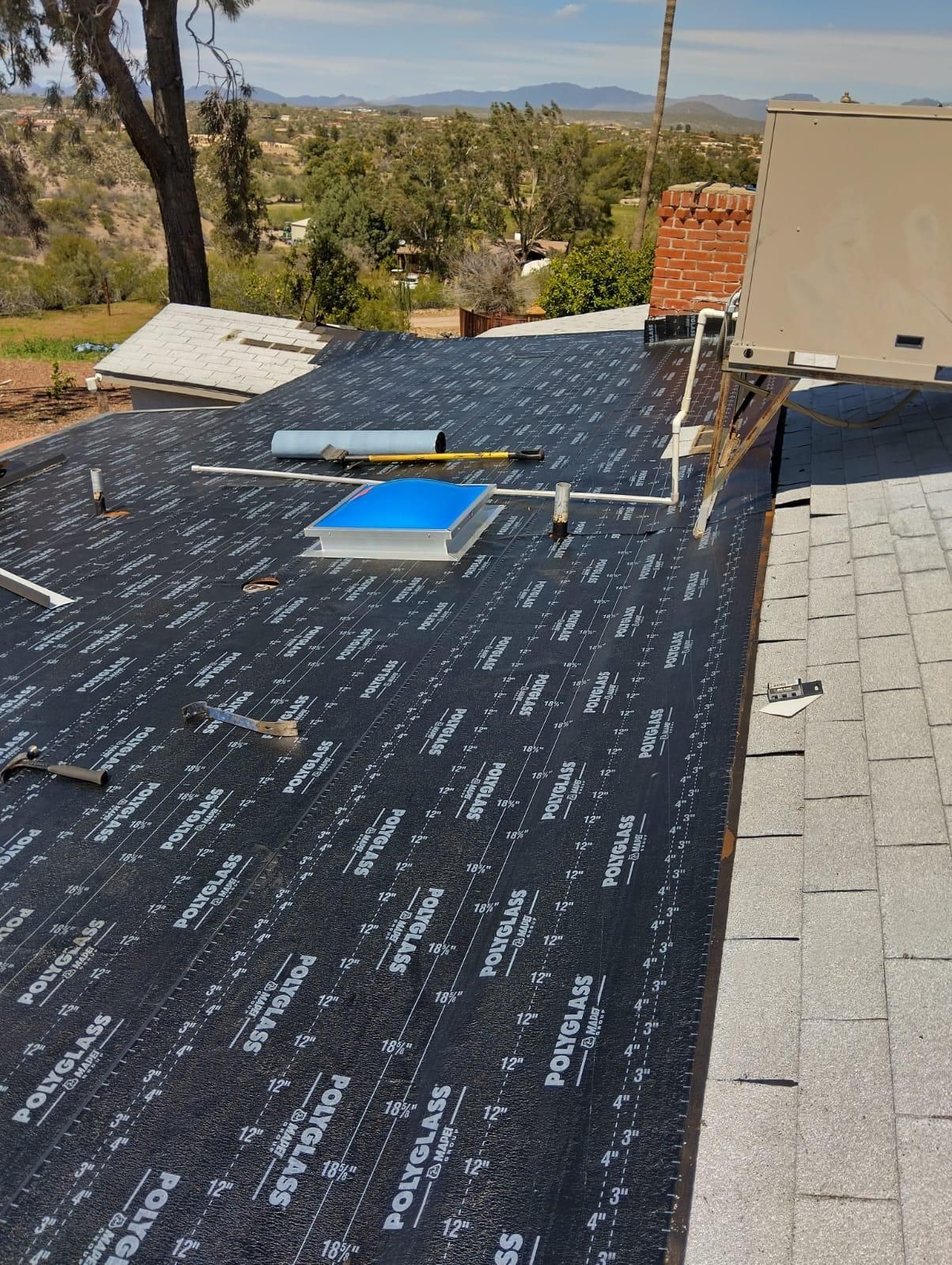 A flat roof with a skylight and asphalt underlayment, viewed on a sunny day with distant hills.
