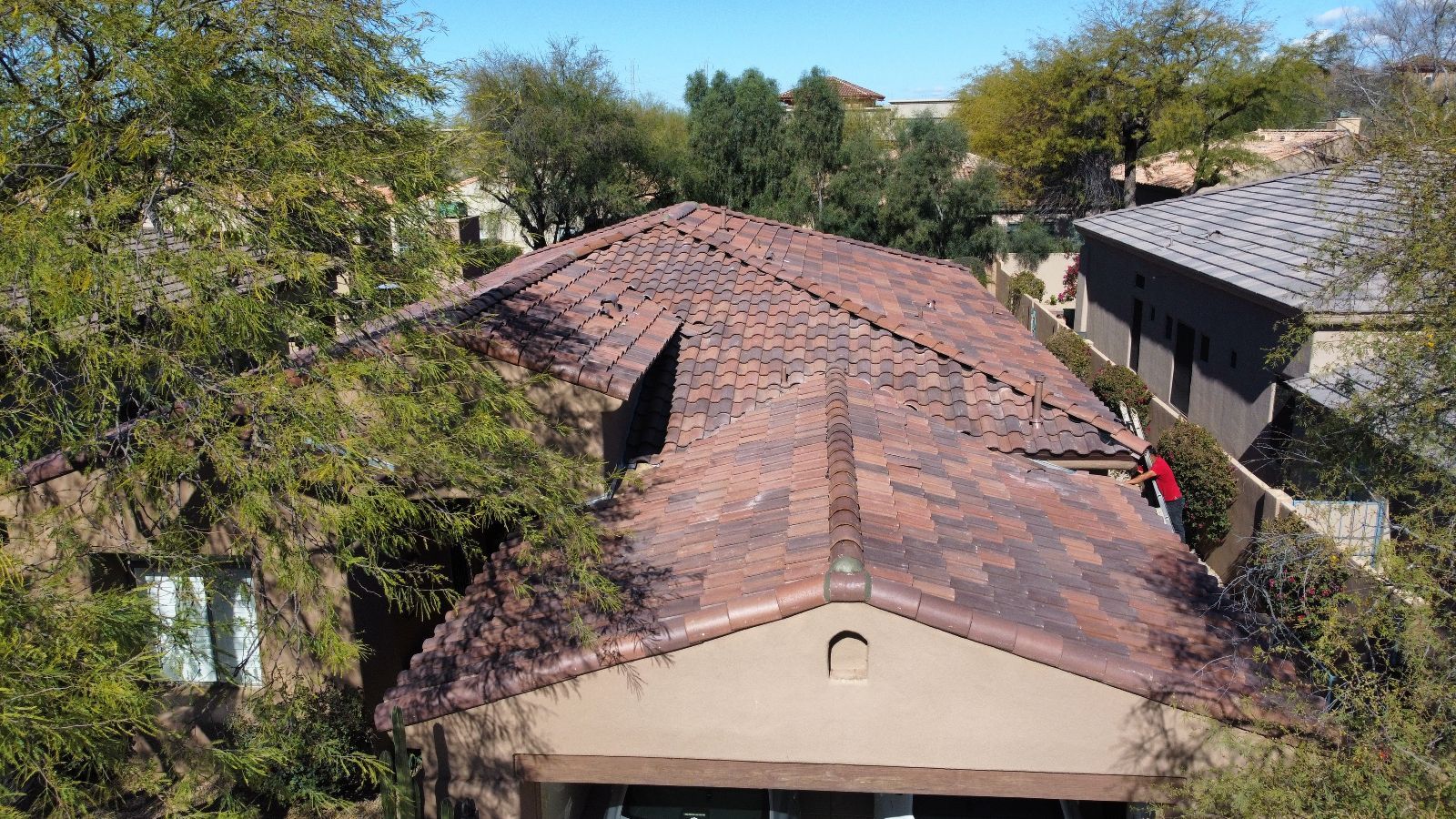 Brown tile roof on a house, viewed from above, surrounded by green trees under a blue sky.