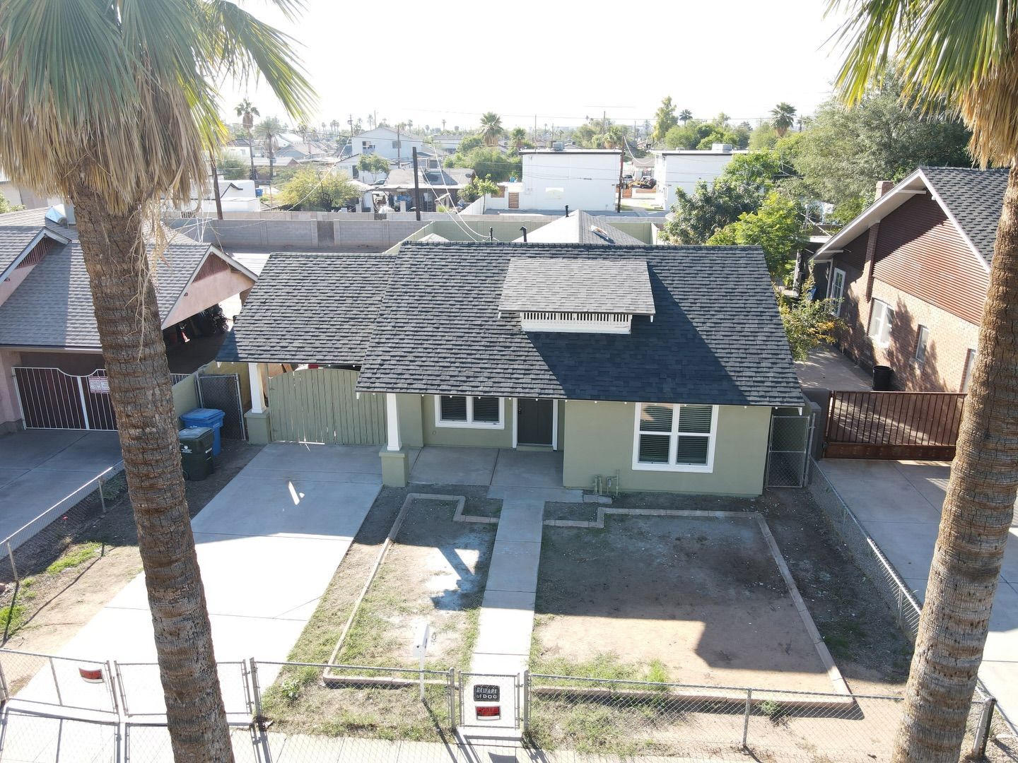 Aerial view of a light green house with gray roof, front yard with dirt patches, and palm trees framing the scene.