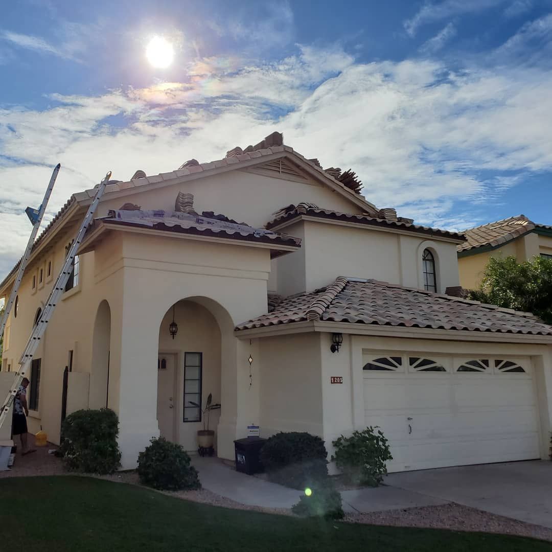 Two-story beige house with red tile roof, arched entryway, garage, and ladder. Sunny day with blue sky.