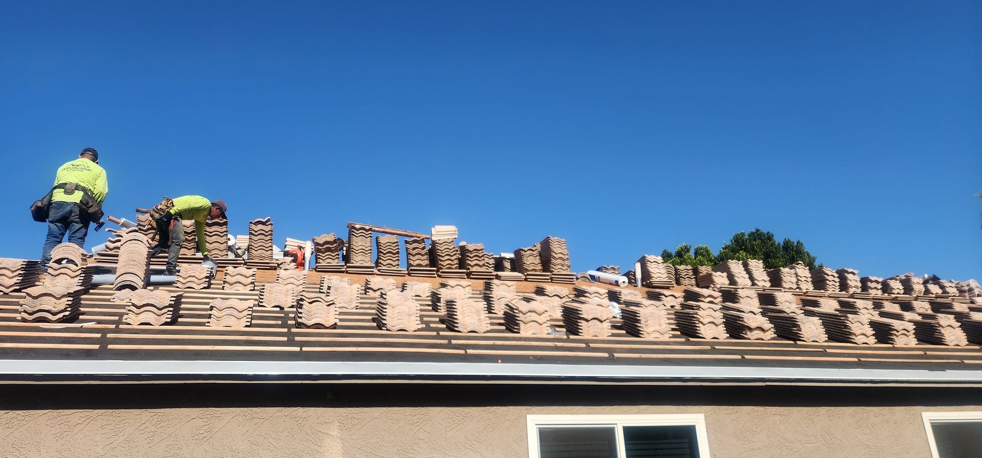 Roofers replacing clay tiles on a house roof under a clear blue sky.