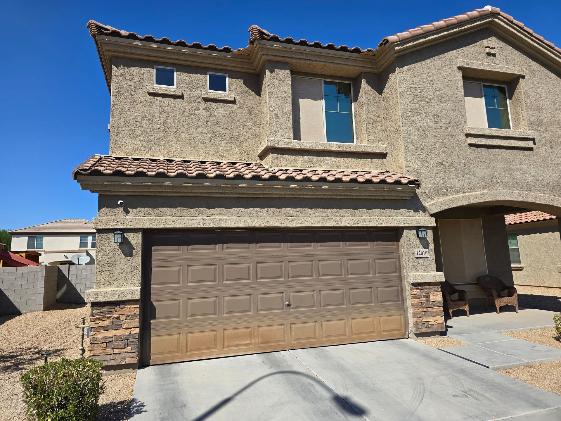 Two-story beige stucco house with brown garage door, tiled roof, and blue sky.