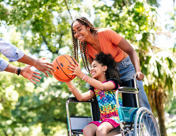 Woman and child in a wheelchair passing a basketball outdoors, smiling with a helper nearby.
