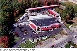Aerial view of a commercial building complex with red and white trim, a large parking lot, and surrounding trees.