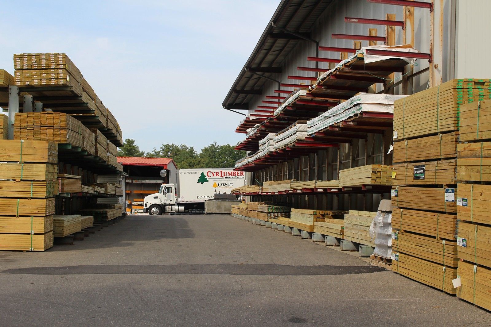 Lumberyard exterior: Stacks of wood planks and sheets, delivery truck in the center.