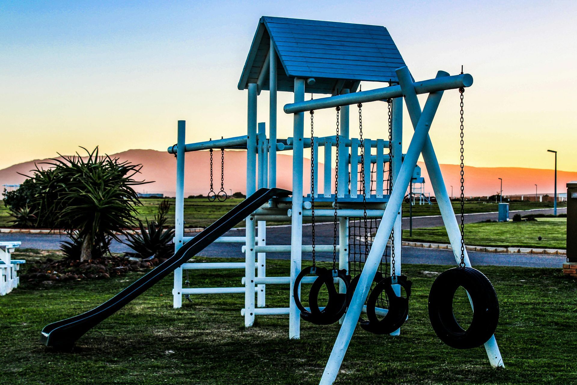 Playground structure with a slide, tire swings, and a small tower against a sunrise backdrop with a mountain range.