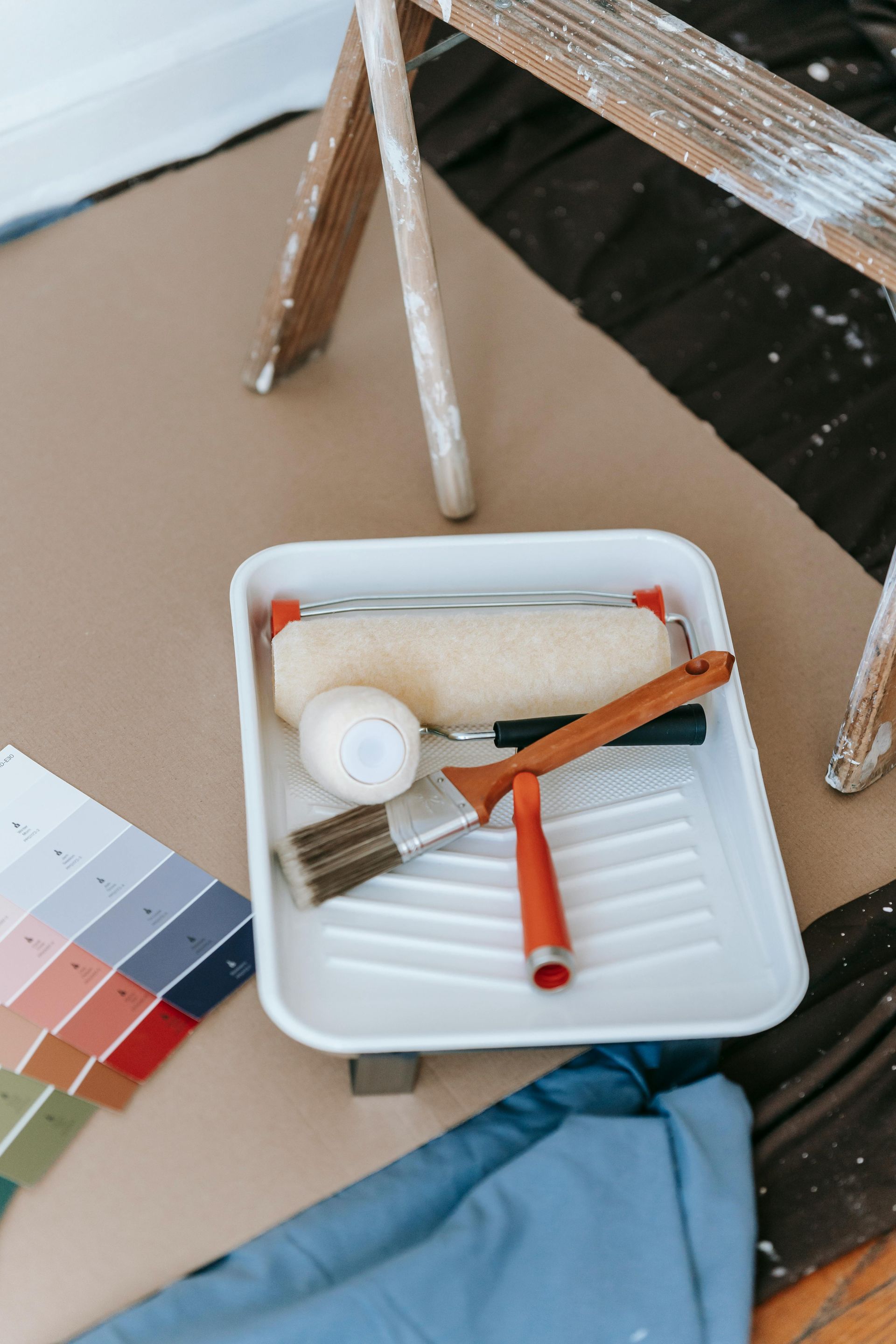 Paint supplies in a tray, including a roller, brush, and color swatches, next to a ladder and drop cloth, preparing for painting.