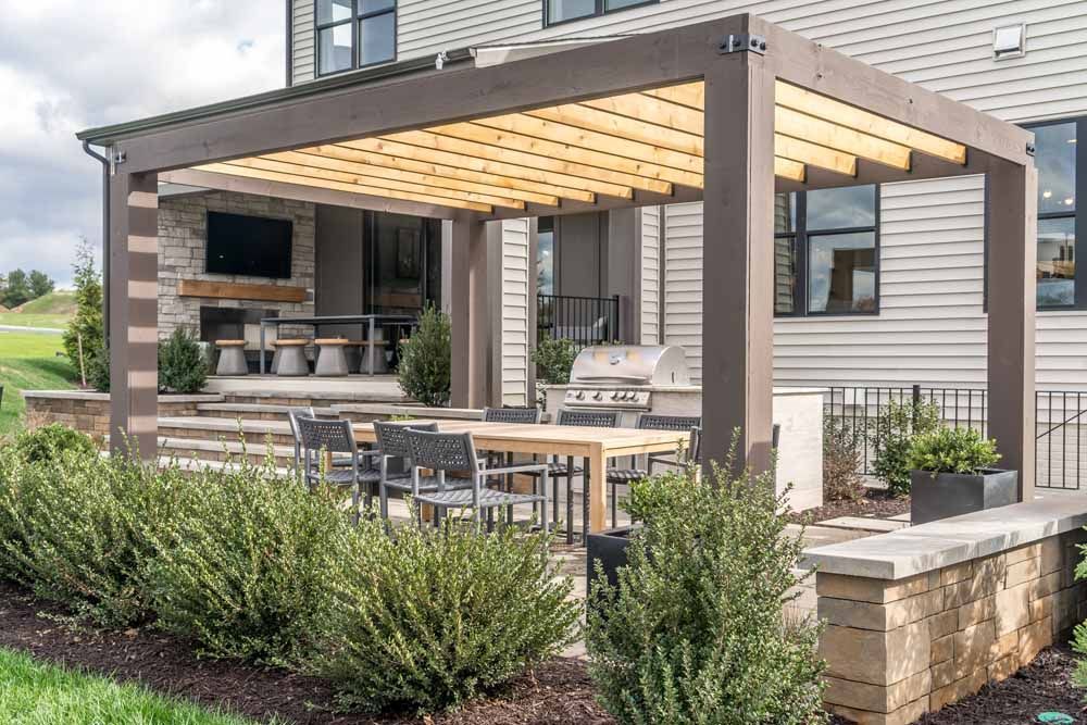 Outdoor patio with a pergola, dining table, and built-in grill. The house has gray siding, and green bushes line the foreground.