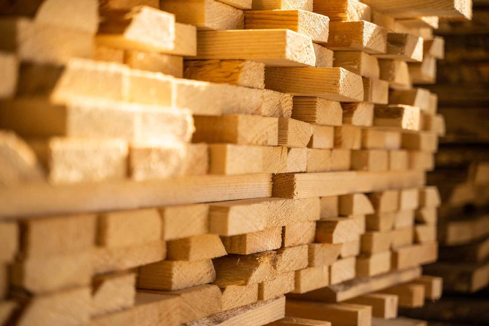 Stack of light-colored lumber planks, tightly arranged in a storage area. The end grains are visible, illuminated by warm light.