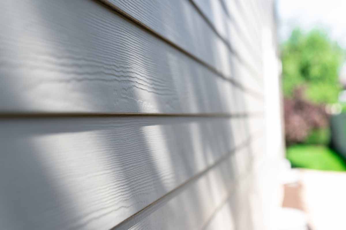 Close-up of light-colored siding on a house, with a blurred green yard in the background.