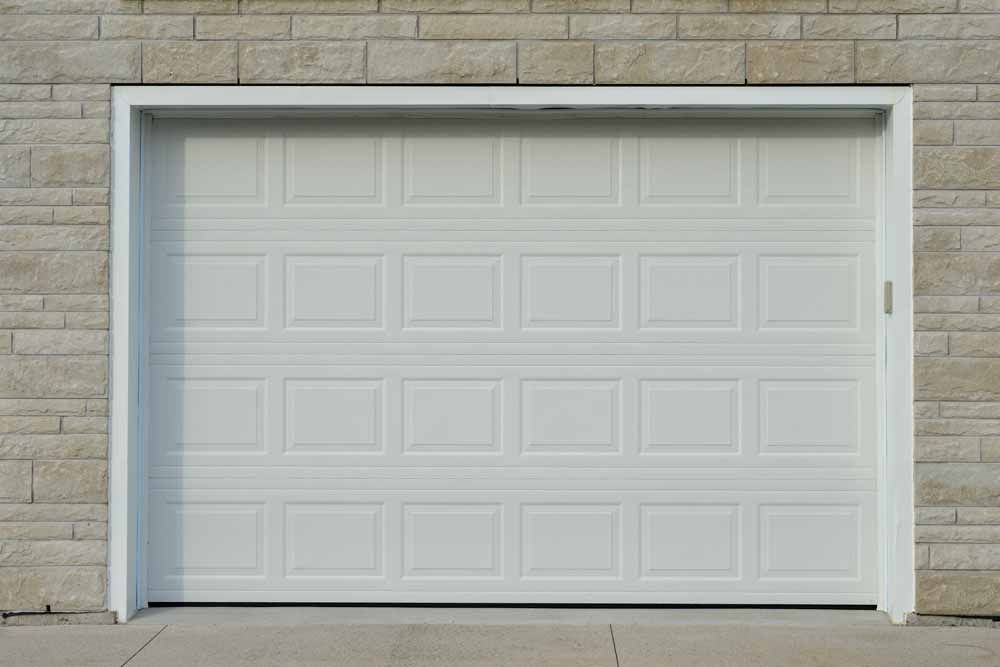 White garage door with rectangular panels, set against a light stone wall, with a concrete driveway.