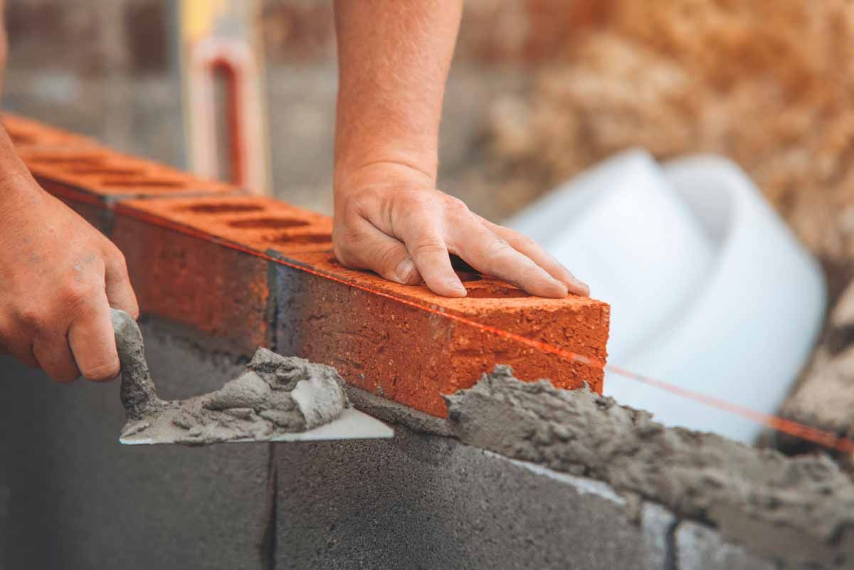 A bricklayer placing a red brick on a concrete block wall, using a trowel to spread mortar.