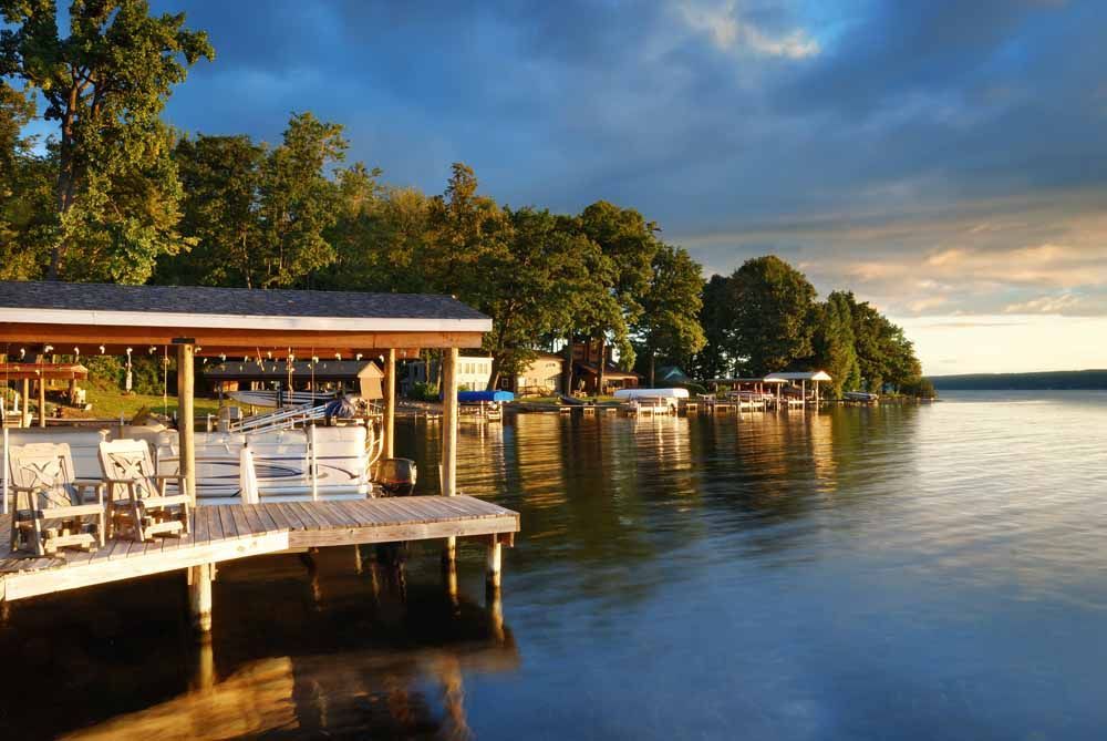 Wooden dock with seating extends into calm lake, lined with trees and docks, under a partly cloudy sky at sunset.