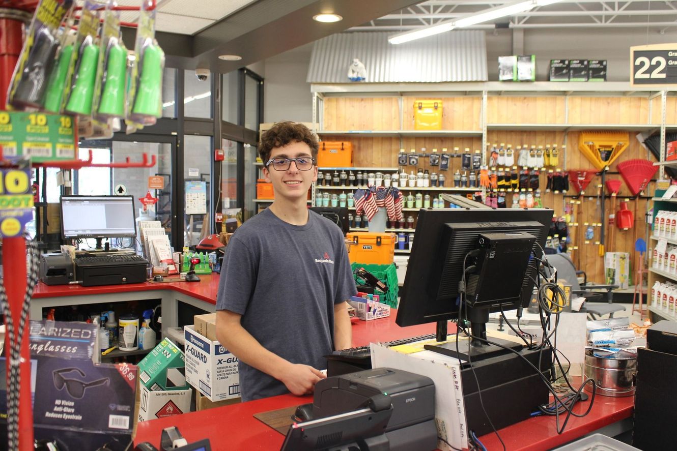 A young man with glasses smiles at a cashier counter in a hardware store. Shelves and tools fill the background.