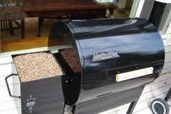 Black pellet grill with open hopper filled with wood pellets.  Grill is on a patio, beside a window.