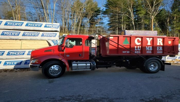 Red Cyr Lumber dump truck parked next to stacks of lumber; outdoors on a paved area with trees in the background.