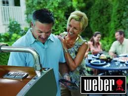 Man grilling on a Weber grill outdoors with woman smiling and putting her hand on his shoulder, other people at table, Weber logo.