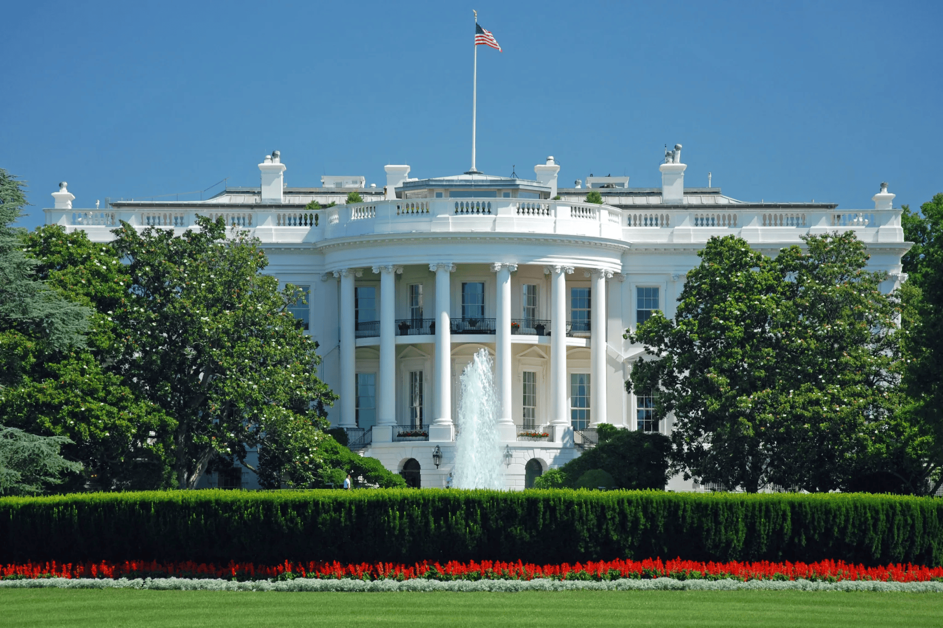 White House, Washington D.C., with a central fountain, green lawn, red flowers, and a blue sky.