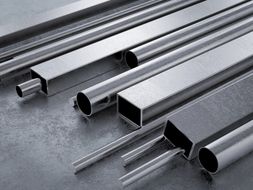 Vari tubi e barre metalliche di diverse forme, tra cui rotonde, quadrate e rettangolari, su una superficie grigia.