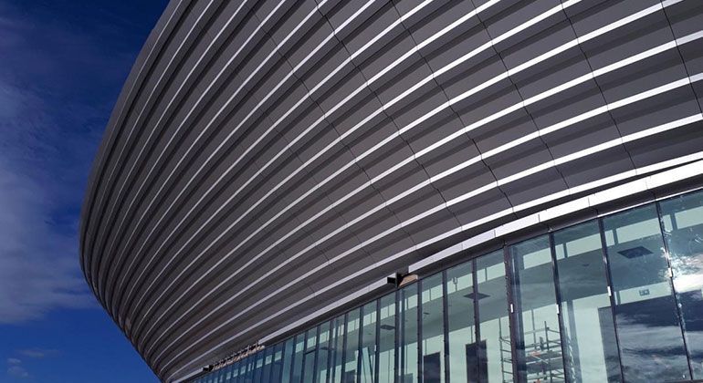 Facciata metallica curva e nervata di un edificio moderno contro un cielo azzurro, sopra una struttura con pareti in vetro.