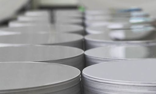 Pile di dischi rotondi di metallo argentato, probabilmente coperchi o pezzi grezzi, in un ambiente industriale.