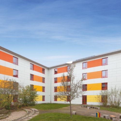 Edificio con strisce colorate arancioni e gialle; cortile con prato e un albero. Cielo azzurro.