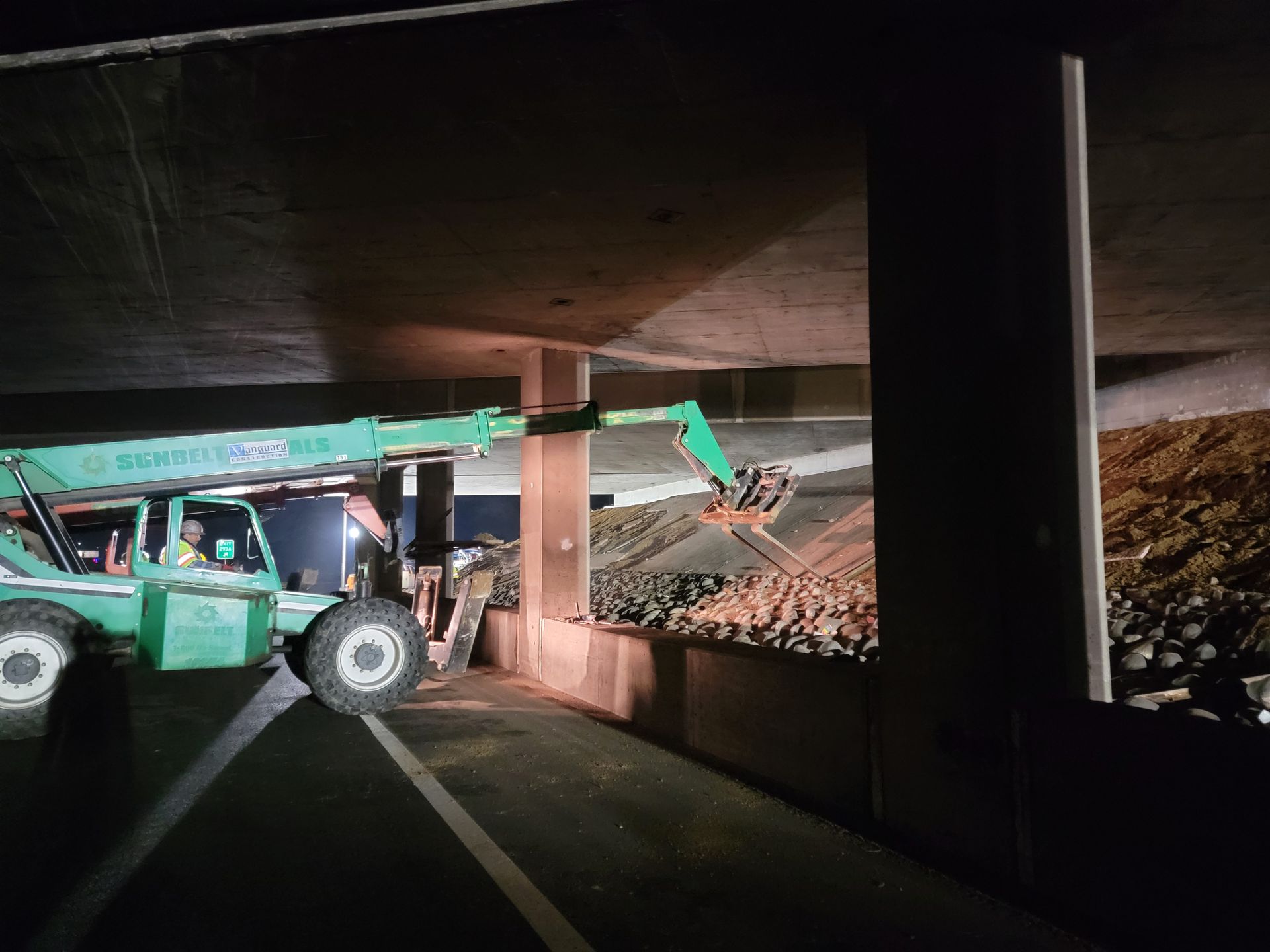 A green forklift is parked under a bridge at night.