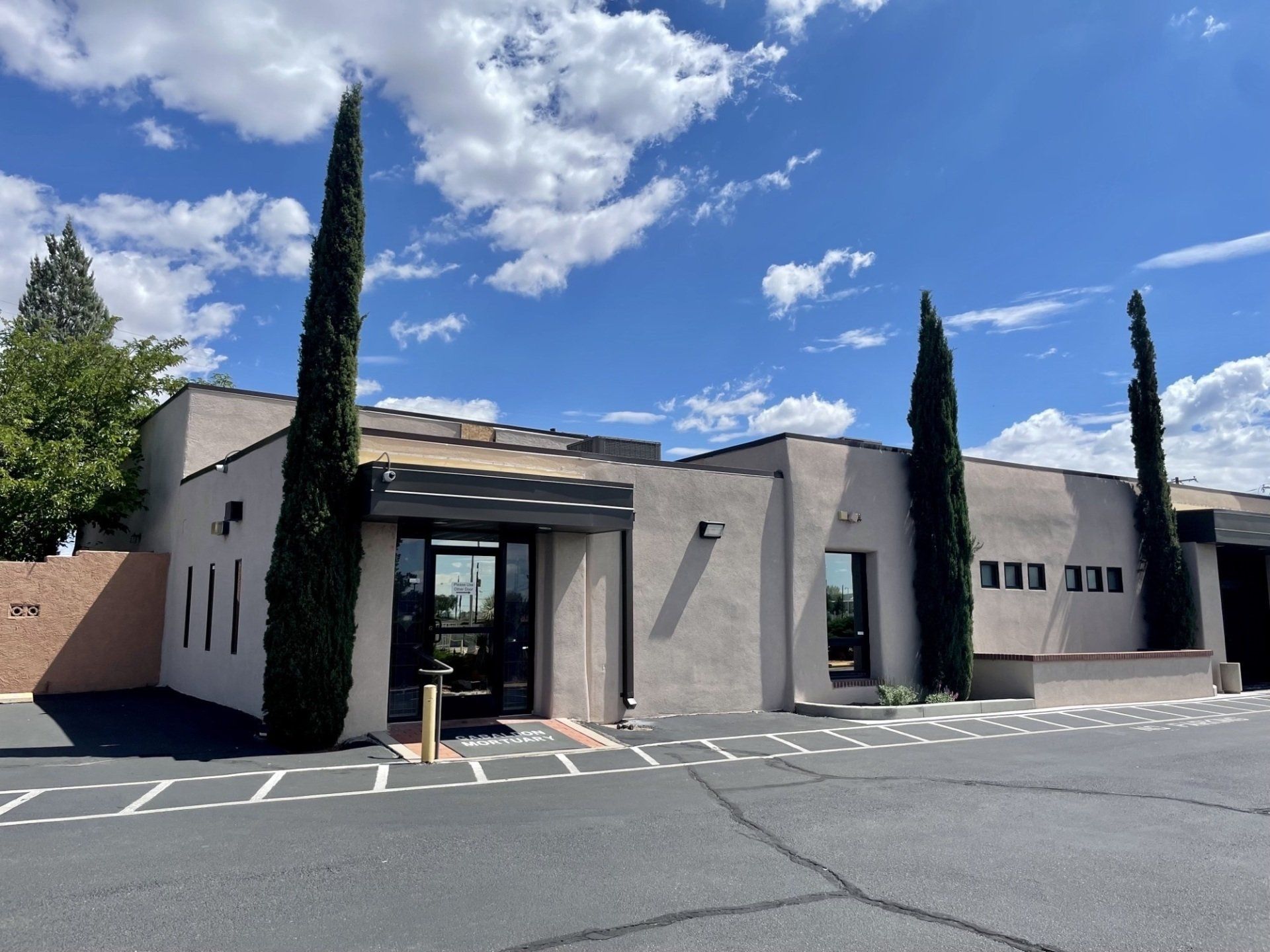 A modern, low-slung building with a dark-trimmed entrance, tall, thin trees, and blue sky.